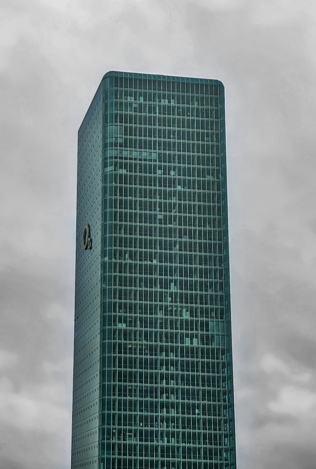 This image showcases the O2 Tower in Munich, Germany, a prominent skyscraper with a sleek glass facade. The high-rise office building stands tall against a cloudy sky, its reflective surface and geometric lines creating a striking contrast with the overcast background. The structures modern architectural design emphasizes its corporate and urban character, making it an iconic part of the citys skyline.