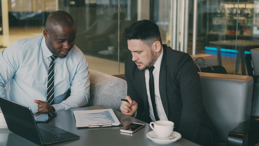 digital Two businessmen in formal clothes sitting and discussing their business financial charts while looking at notepad in glassy modern cafe during lunch time
